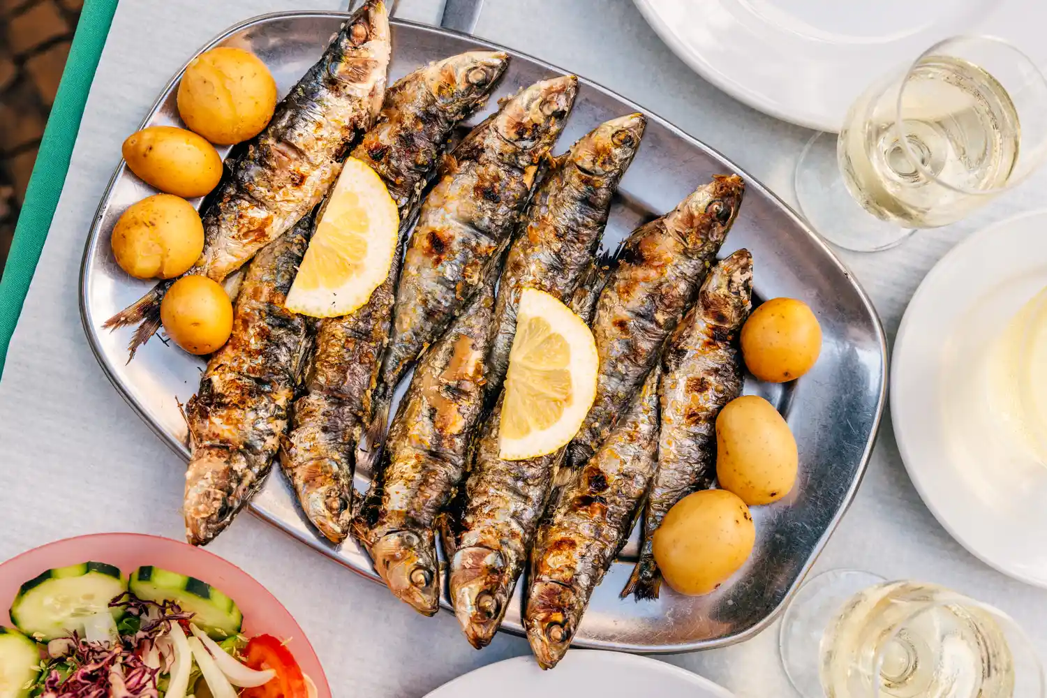 A serving platter with grilled sardines garnished with lemon slices and served with boiled potatoes