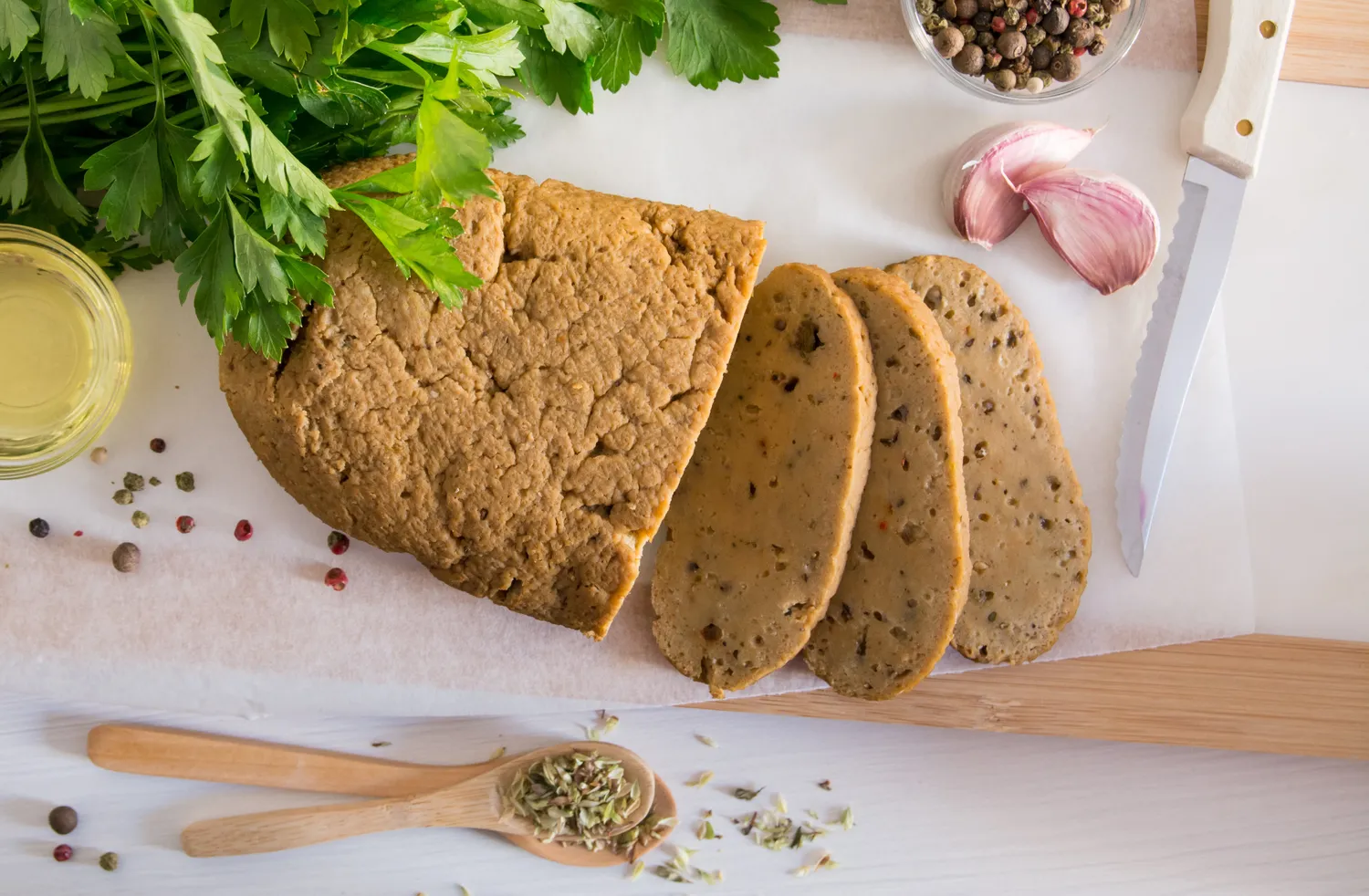 Sliced loaf of seitan on a cutting board with herbs garlic and spices around it