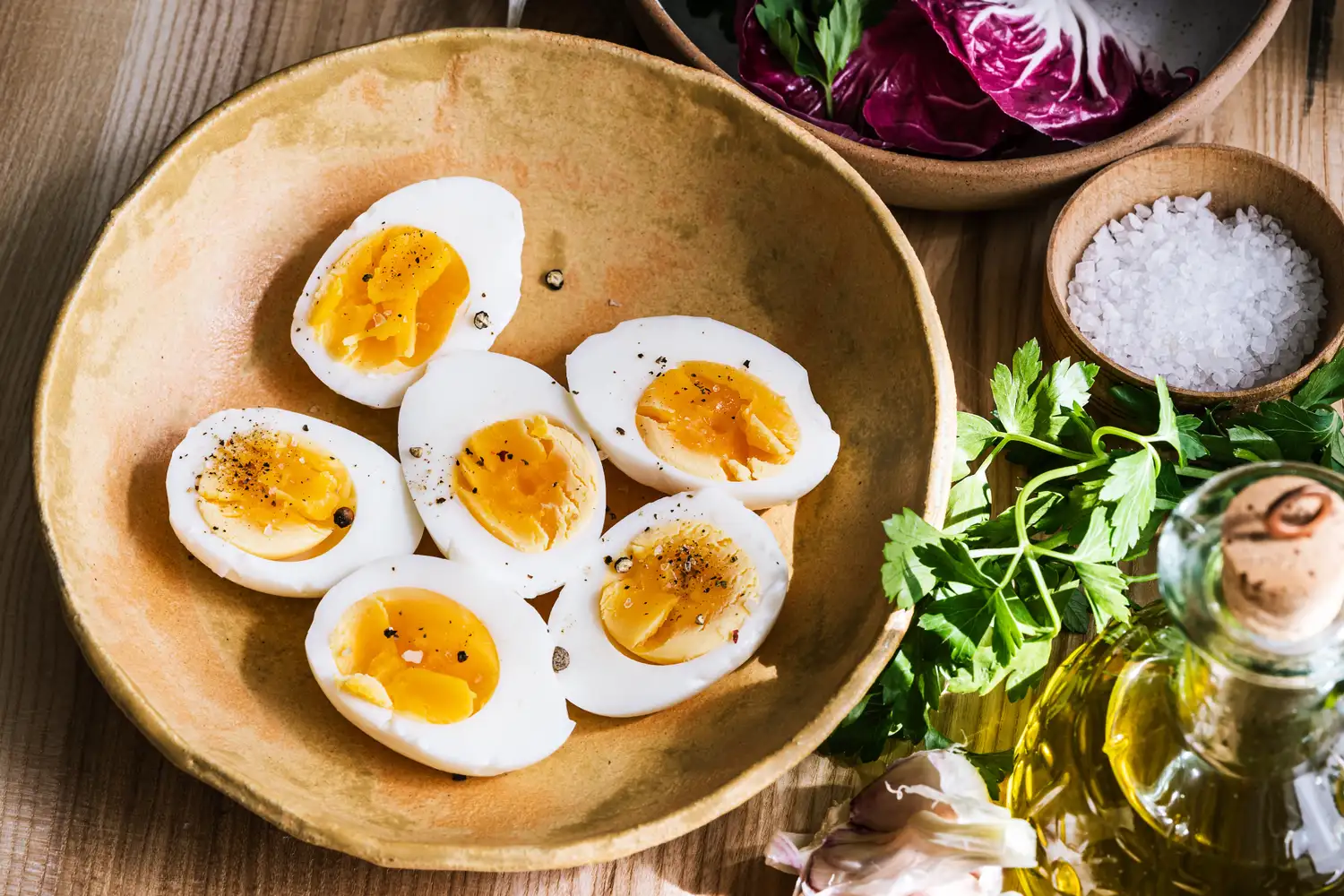 A bowl of sliced boiled eggs topped with ground pepper surrounded by garnishes such as greens radicchio and a bottle of olive oil