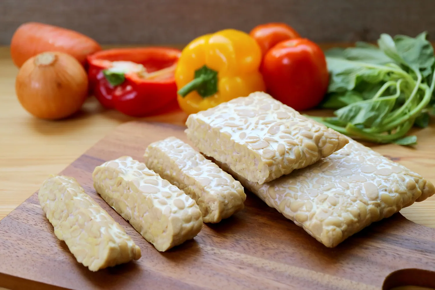 Tempeh blocks on a cutting board surrounded by fresh vegetables including bell peppers onion and greens as background ingredients