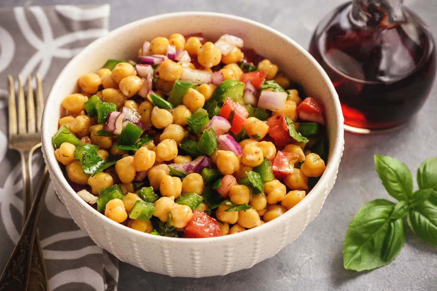 A bowl of chickpea salad with vegetables like tomatoes and greens placed next to basil leaves and a container of dressing